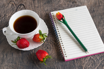 Morning white coffee cup with strawberries on a wooden table with notepad.