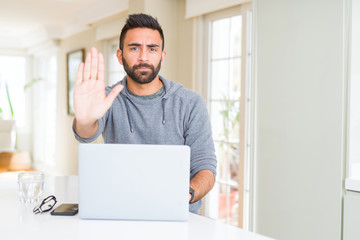 Handsome hispanic man working using computer laptop with open hand doing stop sign with serious and confident expression, defense gesture