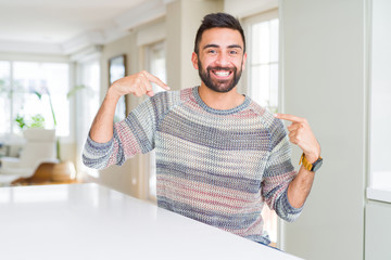 Handsome hispanic man wearing casual sweater at home looking confident with smile on face, pointing...