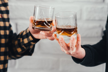 Two hands clink glasses of whiskey on a white brick background