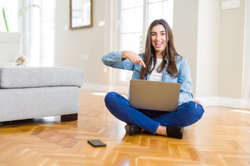 Beautiful young woman sitting on the floor with crossed legs using laptop very happy pointing with hand and finger