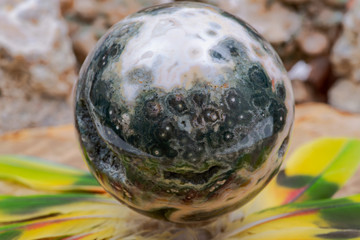 Orbicular Ocean Jasper sphere with crystallized vugs from Madagascar in the middle of a circle made of colorful parrot feathers.