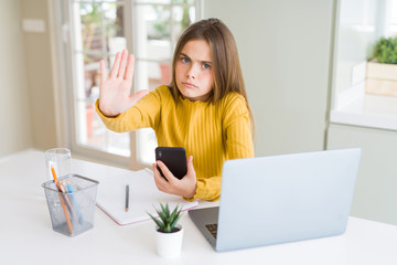 Beautiful young girl kid using smartphone and computer laptop with open hand doing stop sign with...