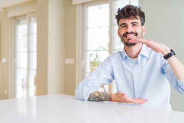 Young businesss man sitting on white table gesturing with hands showing big and large size sign, measure symbol. Smiling looking at the camera. Measuring concept.