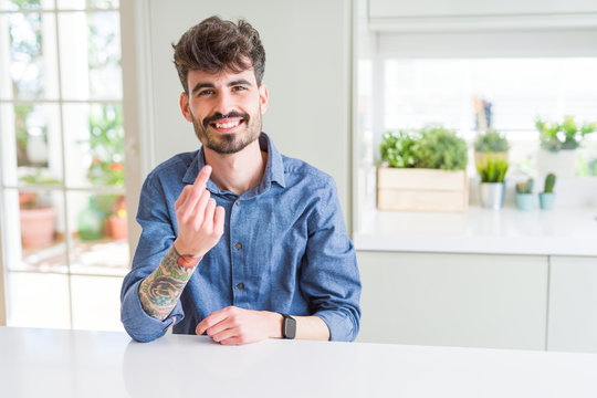 Young man wearing casual shirt sitting on white table Beckoning come here gesture with hand inviting happy and smiling
