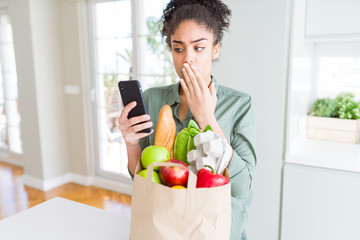 Young african american girl holding paper bag of groceries and using smartphone cover mouth with hand shocked with shame for mistake, expression of fear, scared in silence, secret concept