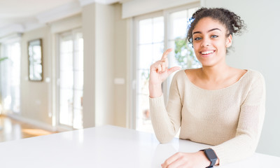 Beautiful young african american woman with afro hair sitting on table at home smiling and confident gesturing with hand doing size sign with fingers while looking and the camera. Measure concept.