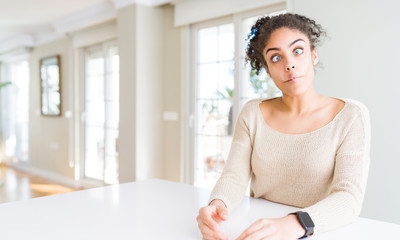 Beautiful young african american woman with afro hair sitting on table at home making fish face with lips, crazy and comical gesture. Funny expression.