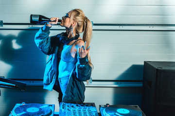 dj woman gesturing near dj mixer while drinking wine from bottle