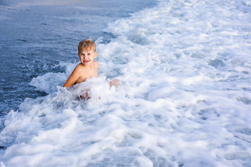 Little blonde child boy siting on sea shore and playing in waves