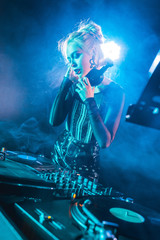 attractive dj girl standing near dj mixer and touching headphones in nightclub with smoke