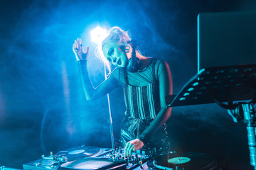 smiling blonde dj girl in headphones standing near dj mixer in nightclub with smoke
