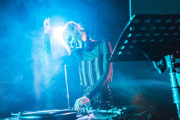 selective focus of happy dj girl in headphones standing near dj mixer in nightclub with smoke - Powered by Adobe