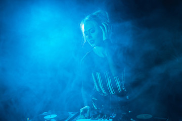 blonde dj girl listening music in headphones near dj equipment in nightclub with smoke