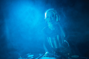 attractive dj girl listening music in headphones near dj equipment in nightclub with smoke