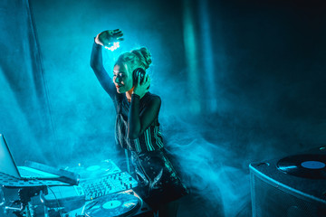 happy dj woman with blonde hair listening music in headphones in nightclub with smoke - obrazy, fototapety, plakaty