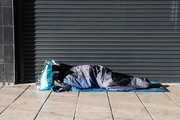 Homeless person asleep in a sleeping bag on a pavement sidewalk in front of a metal shutter.  Face...