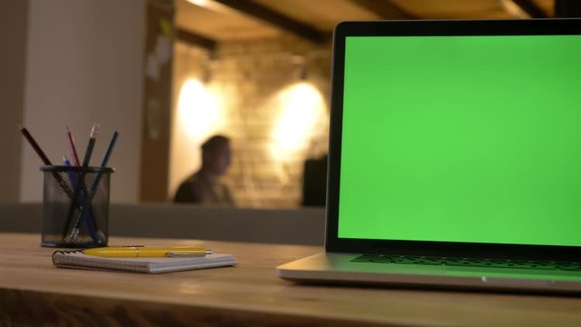 Closeup shoot of green screen of the laptop lying on the desk next to the cup with pencils indoors in the office with an employee on the background