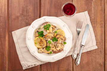 Scallopine di vitello, a traditional Italian dish of veal, with a mushroom sauce, lemons, and parsley, shot from above on a dark rustic wooden background with wine