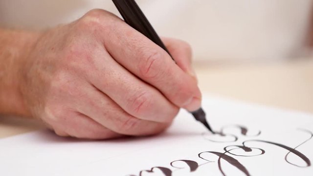 Calligraphist drawing letters on white blank. Close shot. Front view. Calligraphic lettering. Mans hand writing on white blank. Designer at work. Selective soft focus. Blurred background
