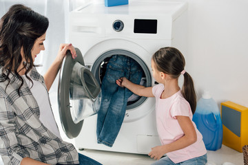 daughter in pink t-shirt and mother putting clothes in washer in laundry room