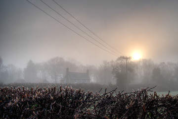 Looking over a headge towards a farmhouse shrouded in mist.  Telegraph poles lead into the scene and the sum.