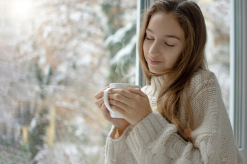 Young woman enjoys drinking hot drink by the window