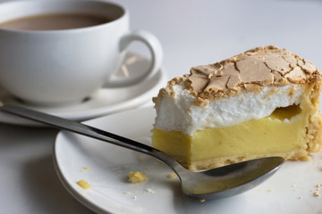 Lemon meringue pie on a small plate with a spoon and cup of tea in the background.  Afternoon tea, snack, teatime, dessert concepts.