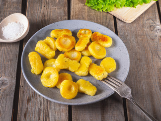 Fried potato gnocchi on a gray ceramic plate, leaves of fresh leaf lettuce lie on an unpainted kitchen board on an old plank rustic table. Next to the plate is a wooden spoon with coarse salt.