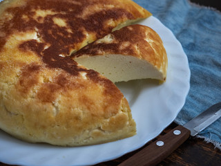 A round curd casserole with a slit lies on a white plate on a stool, next to a denim cloth and a kitchen knife, shot from close range.