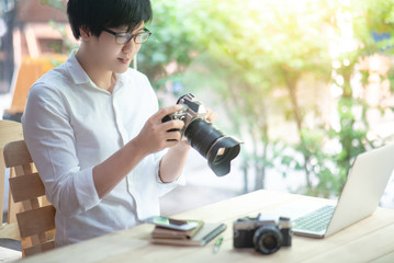 Young Asian man holding digital single-lens reflex camera (DSLR) checking photo on camera screen display sitting with laptop computer and smartphone on the table. Photography, art and hobby concepts