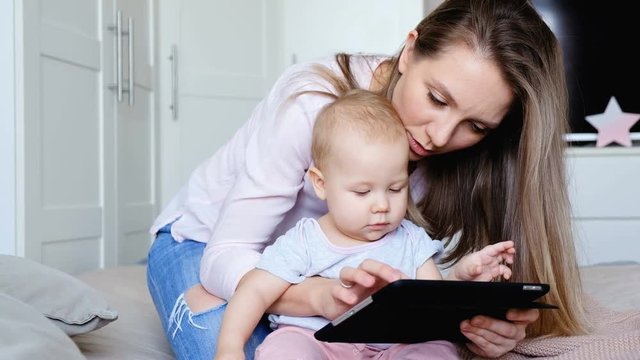 Young positive mother works with her charming baby and shows her developing cartoons on a tablet while sitting on a bed in a cozy bedroom. Concept of decree and child care