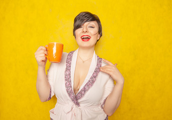 cute woman with short hair dressed in a cozy pink Bathrobe and drinks a tea from an orange Cup on a yellow solid background in the Studio