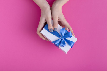 girl holding a beautiful present in hands, women with gift box with a tied blue ribbon bow in hands on a colored pink cardboard background, top view, concept holiday, love and care