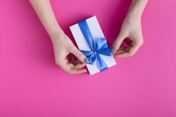 girl holding a beautiful present in hands, women with gift box with a tied blue ribbon bow in hands on a colored pink cardboard background, top view, concept holiday, love and care