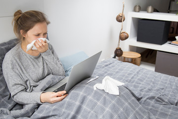 Sick woman working on laptop at home, blowing nose with napkin