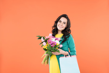 Curly pregnant woman holding flower bouquet and shopping bags isolated on orange