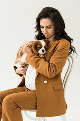 Curly pregnant woman sitting on chair and holding dog isolated on white