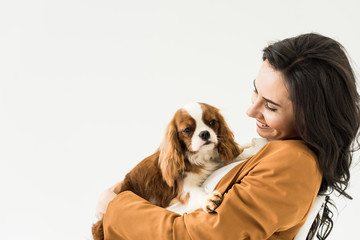 Excited brunette woman in brown jacket holding dog with smile isolated on white