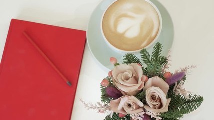 Cappuccino in a cup with a heart on the crema. Coffee with a red notebook and a small bouquet on the table. View from above