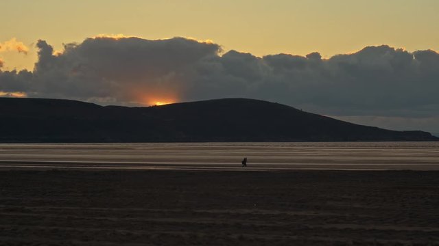 Medium telephoto shot on the beach during a moody cloudy sunset, panning along with the silhouette of a man as he walks along and stops to set up his tripod to photograph the scenery.