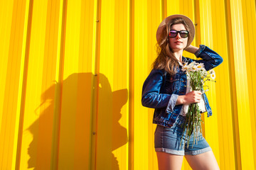 Portrait of hipster girl wearing glasses and hat with flowers against yellow background. Summer outfit. Fashion. Space.