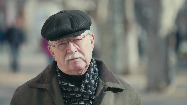 Wise elderly man in a flat cap and glasses standing in a sunny alley in slo-mo   
