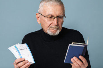 Old pensive man with gray hair and beard in eyeglasses and sweater thoughtfully holding tickets and passports in hands over blue background isolated