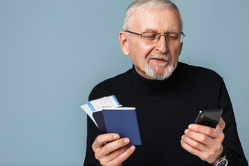 Old attractive man with gray hair and beard in eyeglasses and sweater holding tickets and passports in hand while happily using cellphone over blue background