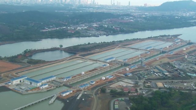 Wide, high aerial view of the massive and improved locks of the Panama Canal near the coast. Modern skyscrapers and the ocean on the horizon. Ships navigating the canal. Drone view.