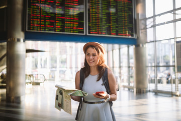 theme travel and transportation. Beautiful young caucasian woman in dress and backpack standing inside train station terminal looking at electronic scoreboard holding phone, map paper hand navigation