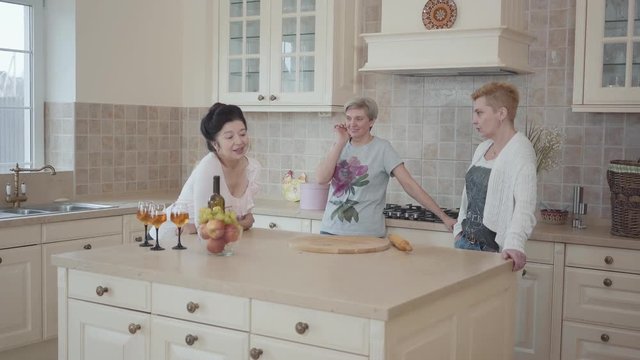 Senior ladies having fun spending time together drinking wine and eating fruits. Three mature women communicating chatting at home standing near modern table in the middle of kitchen.