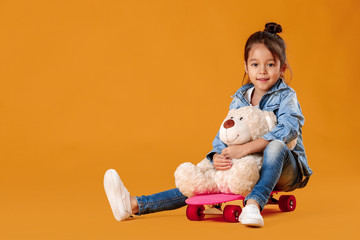 Naklejka premium happy little child girl in jeans clothes sitting with Teddy bear on skateboard on orange background