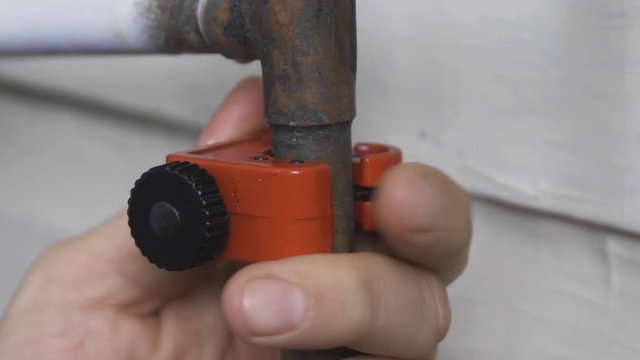 Male hands turn an orange pipe cutter to open an old copper pipe.